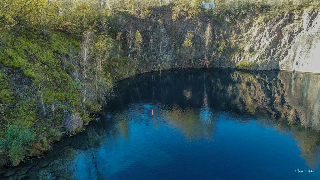 Vue aérienne de la carrière de Vodelée avec eau bleue, falaises et végétation, photo par Maxime HIBLOT